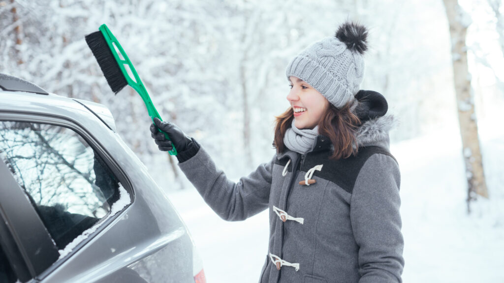 Smiling woman brushing snow off her car with a snow brush during winter.